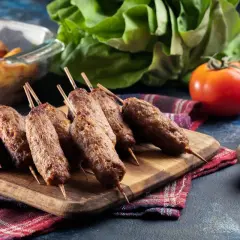 Grillowane ćevapi na drewnianej desce, obok pomidorów, czosnku i pieczonych ziemniaków.