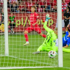 Napięta akcja w polu karnym podczas meczu Widzew Łódź – Lech Poznań. Bramkarz w zielonym stroju broni strzał, a zawodnik w niebieskiej koszulce leży na murawie.