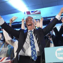 Jubelnde Menge bei einer Wahl in England. Ein Mann mit erhobenen Armen und einem breiten Lachen im Vordergrund, umgeben von Union-Jack-Flaggen und "Reform UK"-Schildern.