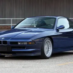 Granatowy Alpina B12 z lat 90. błyszczy na tle industrialnego budynku. Złote detale podkreślają jego sportowy charakter.