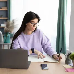 Kobieta w okularach pisze w zeszycie, szukając informacji, jak znaleźć swój numer IBAN. Obok leży laptop i telefon.
