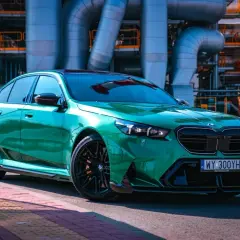 Błyszczące, zielone BMW M5 G90 na tle przemysłowej infrastruktury.