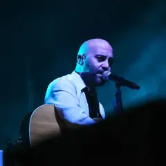 Giuliano Sangiorgi dei Negramaro, con la sua chitarra, canta al microfono, un passo indietro sul palco illuminato da luci blu.