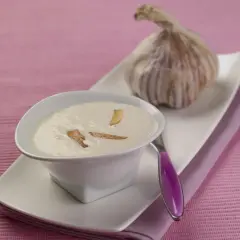 Un bol de crème d'ail onctueuse, garnie de fines lamelles d'ail croustillantes, à côté d'une tête d'ail fraîche.