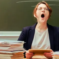 Lehrerin mit Brille gähnt erschöpft hinter einem Stapel Bücher. Ein Klischee, das viele kennen.