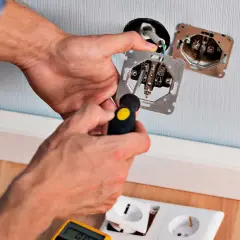 Elektryk montuje gniazdko, sprawdzając, z której strony faza w gniazdku. W rękach trzyma śrubokręt i mechanizm gniazdka.