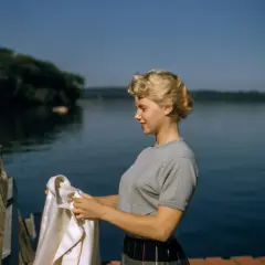 Joven con peinados años 50, con un suéter gris y falda a cuadros, sostiene una chaqueta blanca junto a un lago.
