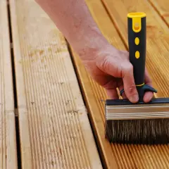 Main d'une personne utilisant une large brosse pour nettoyer bois après ponçage. La terrasse en bois est prête pour le traitement.
