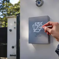 Main MIPA LEFTERD, une main trace un motif géométrique sur une plaque. Une caméra de surveillance est visible au-dessus, comme un marquage cambriolage discret.