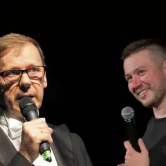 Dwóch mężczyzn z mikrofonami na scenie. Jeden w garniturze, drugi w czarnej koszulce. Widać, że socha stand up.