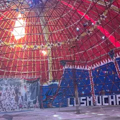 Interior de carpa de circo con techo rojo y azul, telón de fondo de acróbatas y luces que forman "MUCHACHOS". ¡Un espectáculo en Bempota, Ourense!