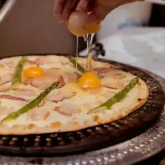 Ręka wylewa jajko na pizzę z boczkiem i szparagami. Pyszna pizza z jajkiem w trakcie przygotowania.