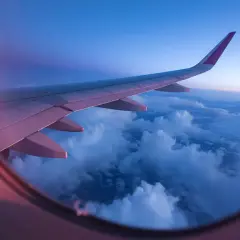 View from an airplane window at sunset. The wing stretches across the frame, bathed in pink and purple light, over a sea of clouds.