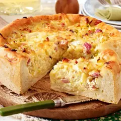 Ein goldbrauner Zwiebelkuchen mit Speck, ein köstliches zwiebelkuchen originalrezept, auf einem Holzbrett mit einem Messer.