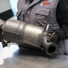 Mechanik w rękawiczkach trzyma metalowy element układu wydechowego samochodu.