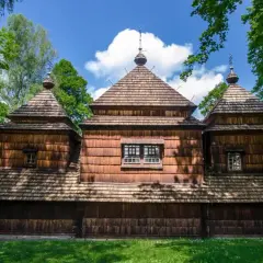 Drewniana cerkiew z charakterystycznymi kopułami, otoczona zielenią. W oddali widać krajobraz, a na pierwszym planie trawa. To miejsce, gdzie czas się zatrzymał, jak w dawnych lutowiskach.