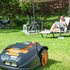 Automatyczna kosiarka do jezdzenia pracuje na trawniku, podczas gdy kobieta odpoczywa na leżaku.
