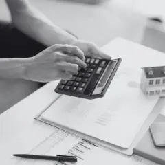 Osoba liczy na kalkulatorze, analizując dokumenty. Obok leży model domu, sugerujący rozliczenie odszkodowania za studzienki na działce.