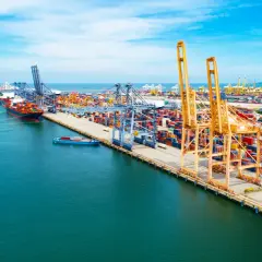 Widok na port kontenerowy z dźwigami, statkami i stosami kontenerów w słoneczny dzień.