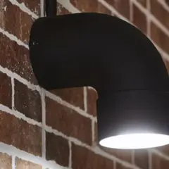 Applique fai da te in metallo nero a forma di gomito, illumina una parete in mattoni rossi.
