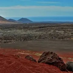 Krajobraz w parku narodowym Timanfaya: czerwone wzgórza, pola lawy i góry na tle oceanu.