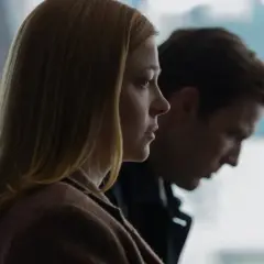 Kobieta z blond włosami i mężczyzna w tle, oboje patrzą w bok. W jej oczach widać smutek, jakby to **all her fault**.