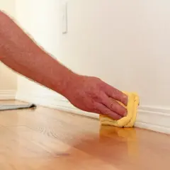 Mano limpiando rodapiés con un paño amarillo. El suelo de madera brilla y la pared está pintada de blanco.