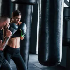 A man with tattoos trains a woman in boxing gloves. They are in a gym with punching bags, showcasing the physical and mental boxing benefits.