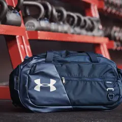 Granatowa torba sportowa 100l z logo Under Armour, gotowa na trening. W tle sztangi i hantle.