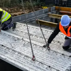 Dwóch budowlańców przygotowuje strop filigran do montażu. Ciężkie płyty zbrojeniowe są podnoszone przez dźwig.