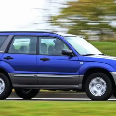 Niebieskie auto 4x4 Subaru Forester z napędem na cztery koła pędzi po drodze, otoczone zielenią.