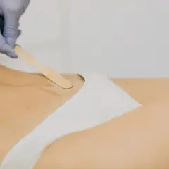 Dłoń w rękawiczce nakłada wosk szpatułką na skórę w okolicach bikini.