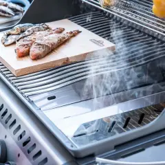 Fisch auf einem Holzbrett auf einem Grill, der zeigt, was ein Smoker Grill kann: Rauch steigt auf und veredelt das Gargut.