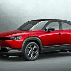 Czerwona Mazda MX-30 z czarnym dachem, stojąca na tle szarej ściany. Stylowy crossover miejski.