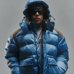 Giovane con piumino blu acceso, cappuccio alzato, occhiali da sole e una collana con ciondolo. Lo sfondo è un grigio chiaro, quasi low red.