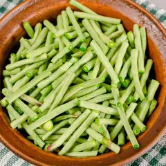 Fagiolini in umido freschi e verdi, pronti per essere cucinati in una ciotola di terracotta su una tovaglia a quadretti.