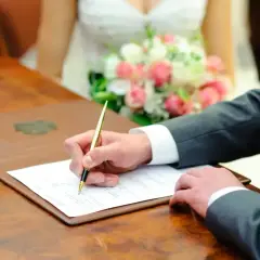 Pan młody podpisuje dokumenty podczas ślubu cywilnego. W tle panna młoda z bukietem róż.