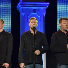 Fünf Männer in schwarzen Hemden singen auf einer Bühne. Sie präsentieren traditionelle kroatische Musik vor einem blauen, pixeligen Hintergrund.