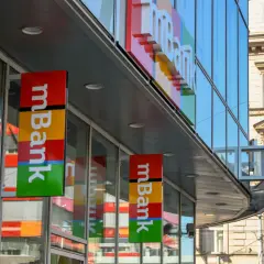 Baner mBank z kolorowym logo i napisem "mBank" na tle nowoczesnego budynku.