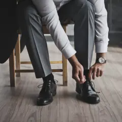 Mężczyzna w garniturze, z marynarką na krześle, zawiązuje buty. Elegancki, śniady kolor stroju i klasyczne obuwie.