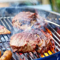 Saftiges Fleisch zum Grillen zischt auf dem Rost. Würstchen und Steaks brutzeln über heißen Kohlen, umgeben von Rauch und Flammen.