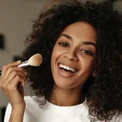 Donna sorridente con capelli ricci applica cipria con un pennello, mostrando come mettere la cipria senza pennello.