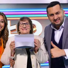Trzy osoby: kobieta w pomarańczowej marynarce, starsza kobieta z kartką i mężczyzna w garniturze pokazujący kciuk w górę.