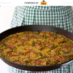 Una persona con un delantal a cuadros verdes sostiene una paella valenciana recién hecha, llena de arroz, pollo, judías verdes y pimientos rojos.