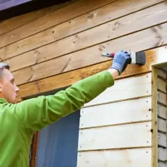 Man målar husfasaden med en pensel. Träet är ljusbrunt och ser ut att vara tryckimpregnerat.