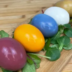 Cinq œufs colorés, du violet à l'orange vif, en passant par le bleu, le blanc et le doré, disposés sur un lit de verdure sur une planche en bois. Chaque couleur oeuf est une surprise.
