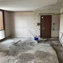 Interno di una stanza in ristrutturazione, con il pavimento in cemento grezzo e pareti spoglie. Si notano attrezzi da lavoro, tra cui un secchio blu e una scopa, che suggeriscono i lavori in corso e i possibili svantaggi dell'intonaco armato.