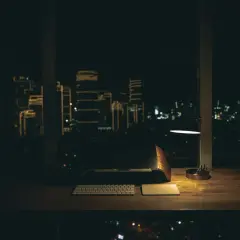 Biurko z laptopem, klawiaturą i lampką nocną. Za oknem nocne miasto, idealne do pracy w godzinach nocnych.