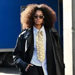 Une femme porte un pantalon noir, une chemise à rayures, une cravate fantaisie et un long manteau en cuir. Ses bottines sont cachées sous son pantalon.