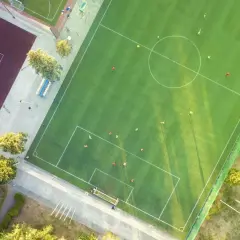 Piłkarze ćwiczą na boisku. Widać całą długość boiska, z liniami, bramkami i zawodnikami w akcji. Obok kort tenisowy.